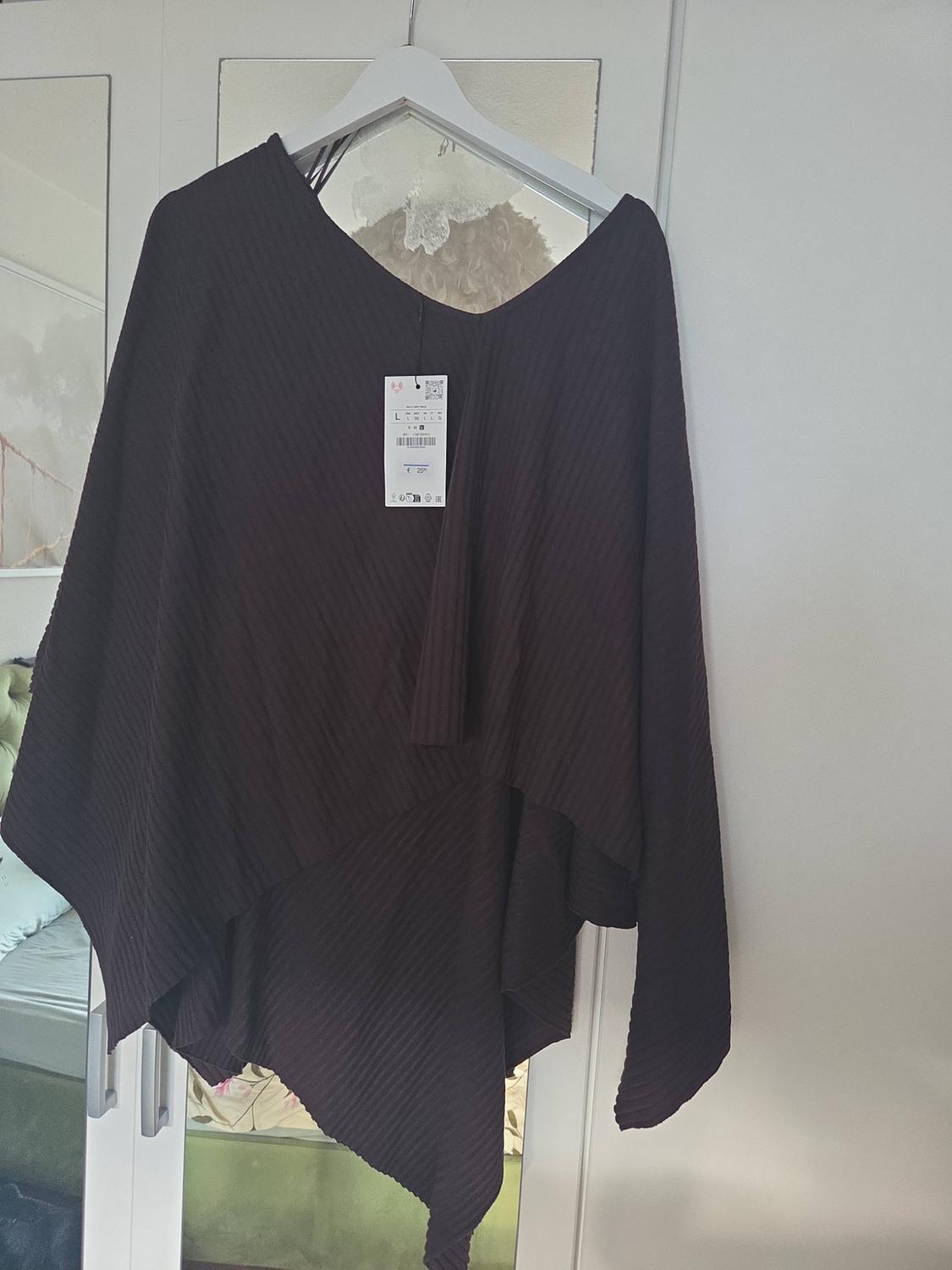 Dark brown asymmetrical ribbed knitted top hanging on a white hanger in front of a mirror. Visible label with the text L and €25.