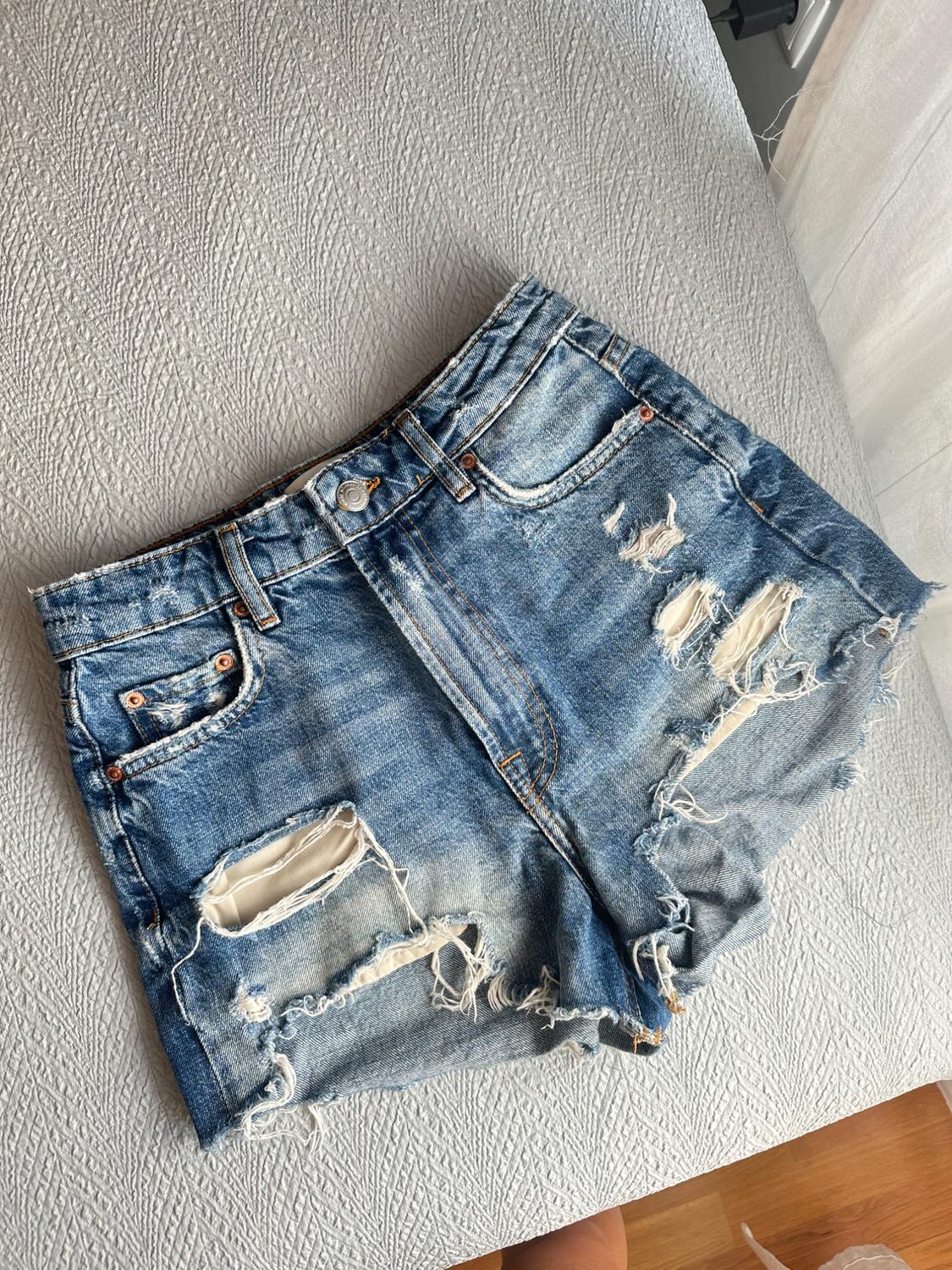 Blue denim shorts with rips revealing the pockets and a frayed hem, on a grey quilt. Casual and relaxed style.