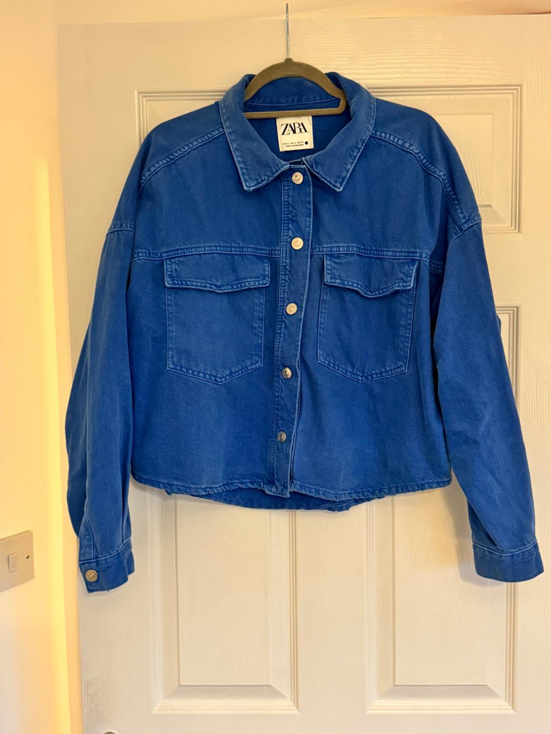 Blue denim jacket with front pockets and metal buttons, hanging on a coat hanger in front of a white door. Zara label visible.