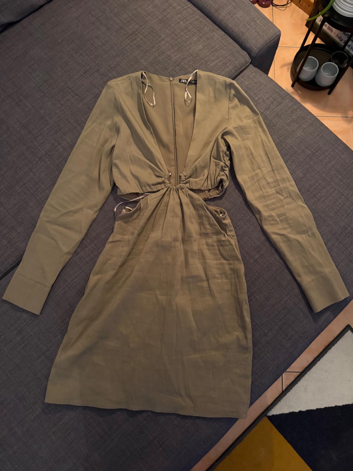 Short, beige dress with long sleeves, a plunging neckline, and ruched detailing at the waist, placed on a grey sofa.