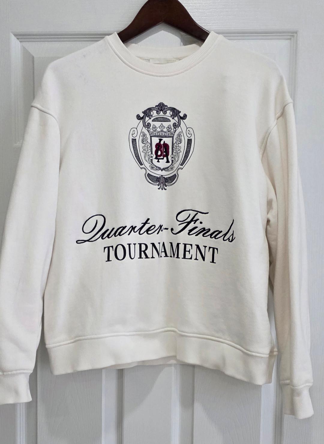 White sweatshirt hanging on a hanger, with a decorative shield and the text "Quarter-Finals TOURNAMENT" on the front. White door background.