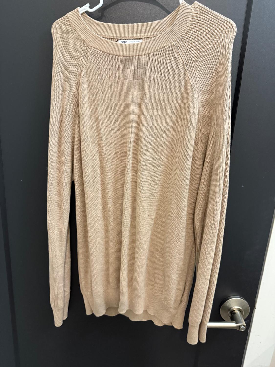 Beige knitted sweater hanging on a white hanger over a black door. Label visible on the neck.