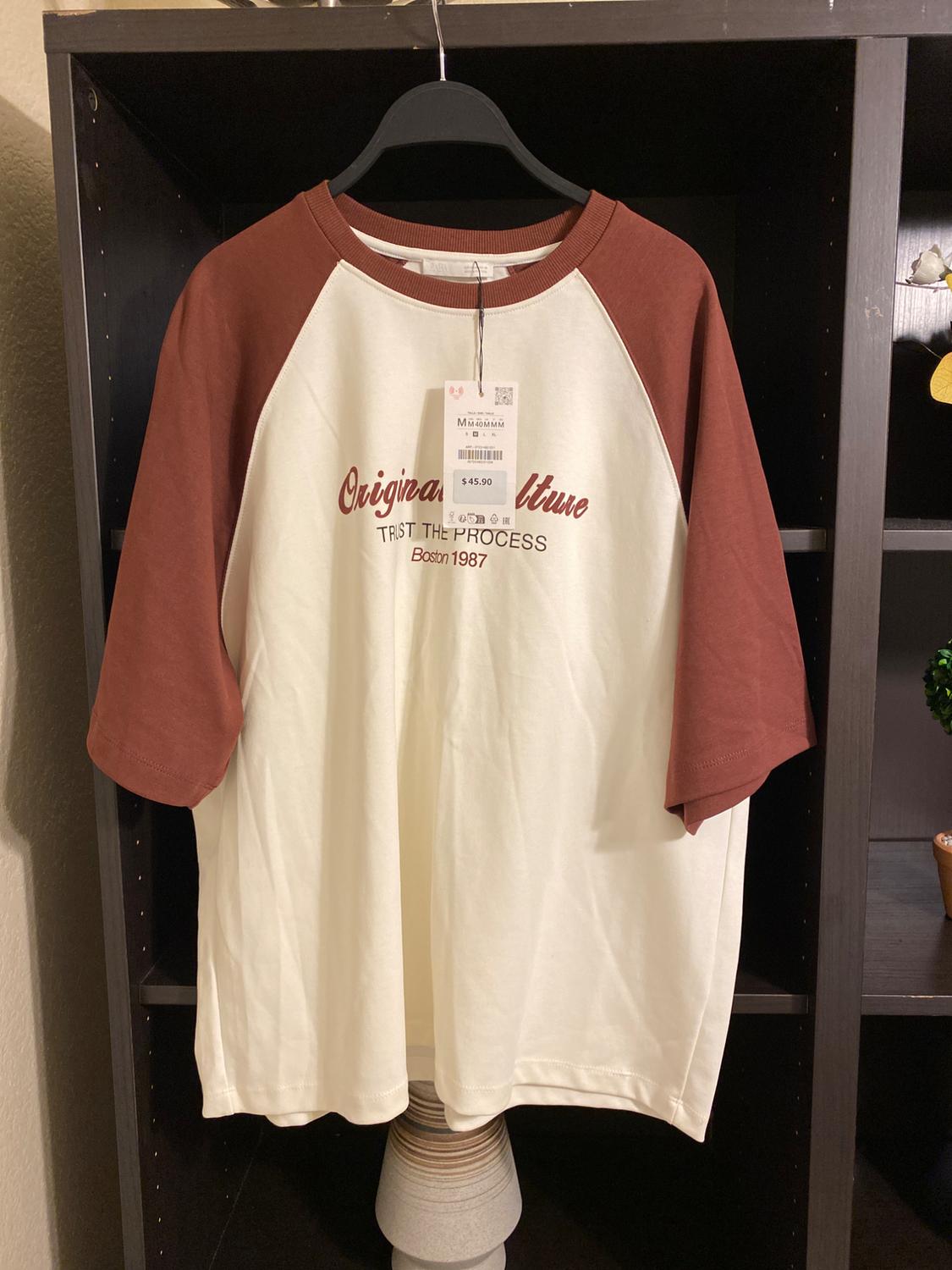 White t-shirt with brown raglan sleeves, text "Original Culture, Trust the Process, Boston 1987", hanging on a black clothes hanger.