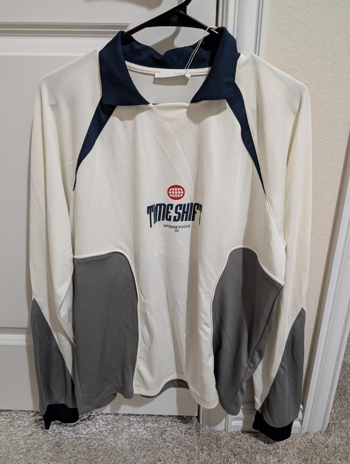 White jumper with grey and navy blue details, hanging on a hanger. Text on the front: "TIME SHIFT INTENSE FOCUS".