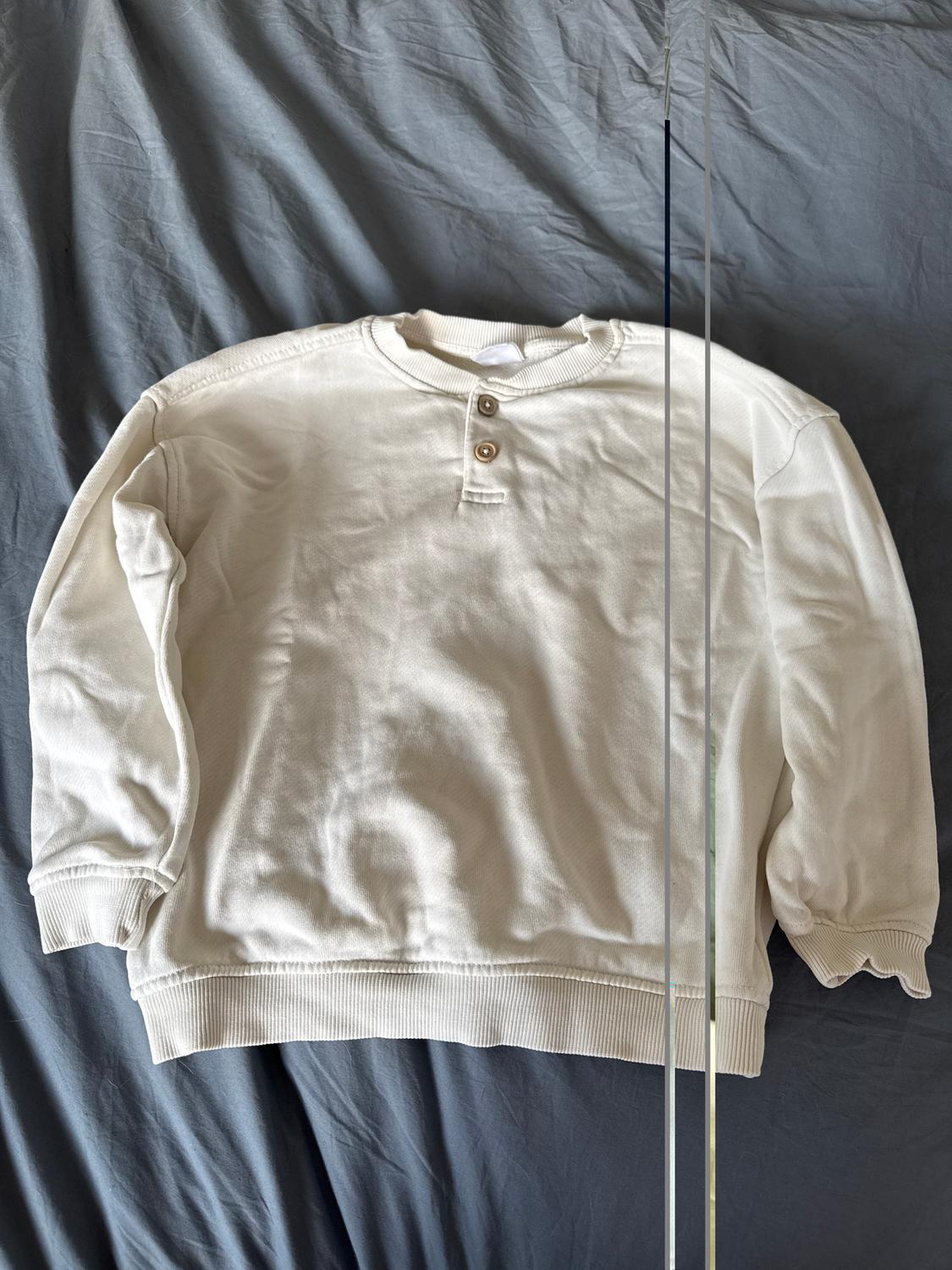 White long-sleeved sweatshirt with buttons on the collar, placed on a wrinkled grey fabric.