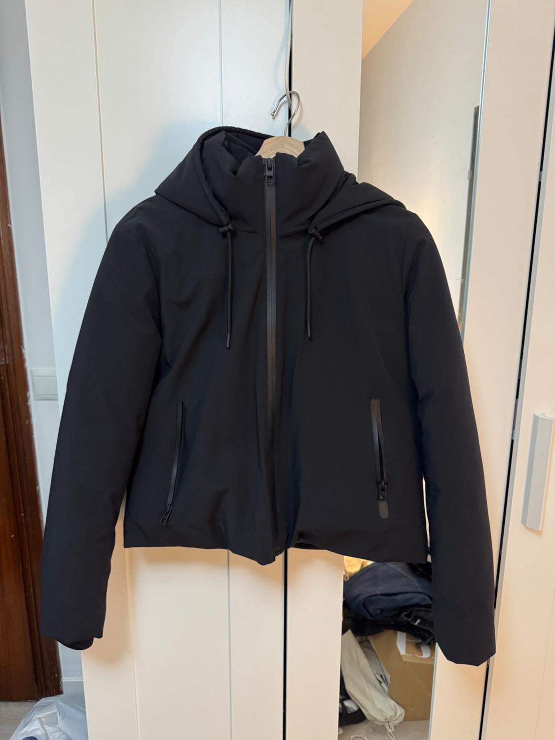 Black hooded jacket with a front zip, hanging on a wooden hanger against a white wardrobe background.