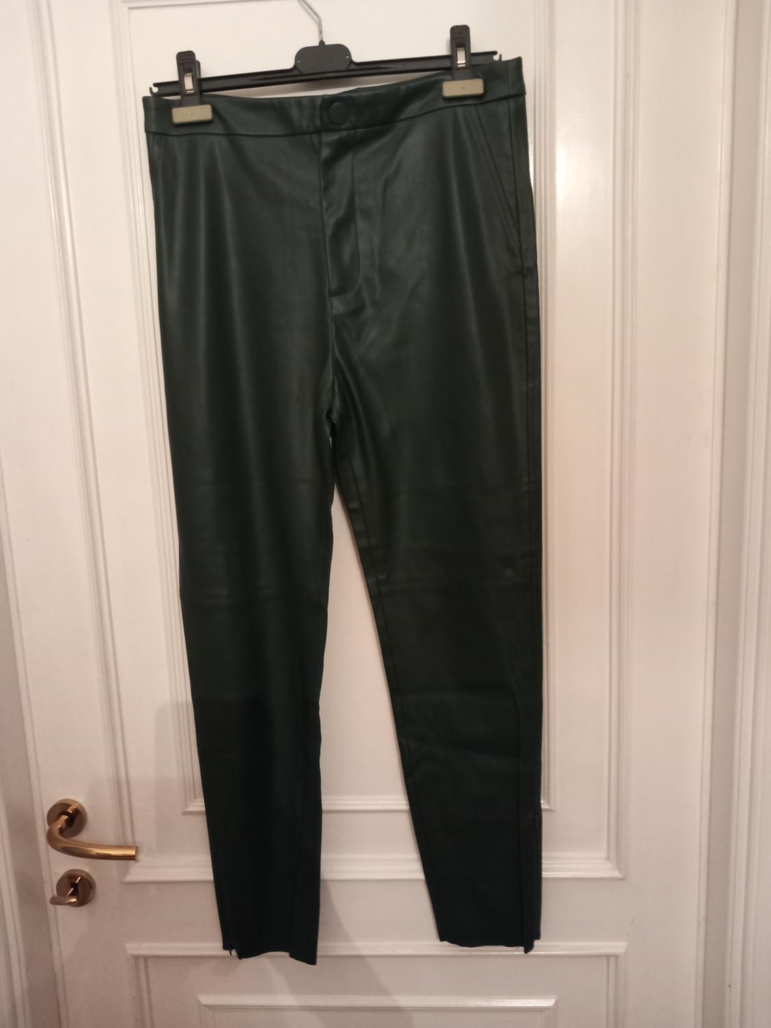 Dark green, glossy-finish trousers hanging on a hanger over a white door with a gold doorknob, in a casual setting.