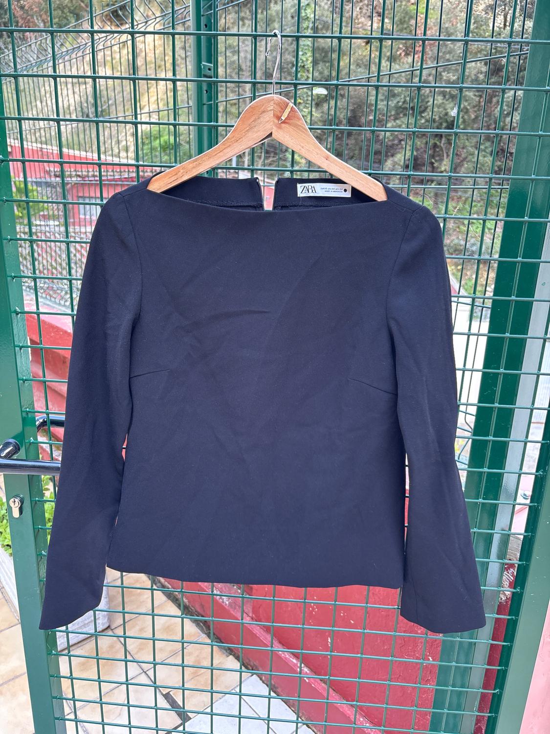 Black long-sleeved blouse hanging on a wooden hanger, green mesh background and natural landscape. Visible tag: Zara.