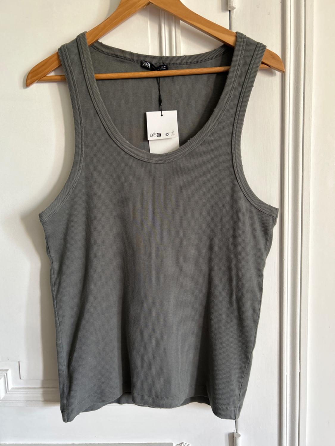 Grey sleeveless t-shirt hanging on a wooden coat hanger, with a visible white label, against a white wall background.