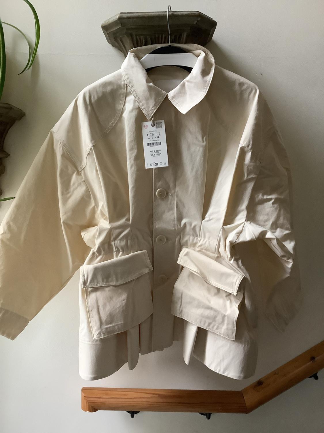 Beige jacket with pockets hanging on a white wall above a wooden banister. Label with text: US $109 UK £109.
