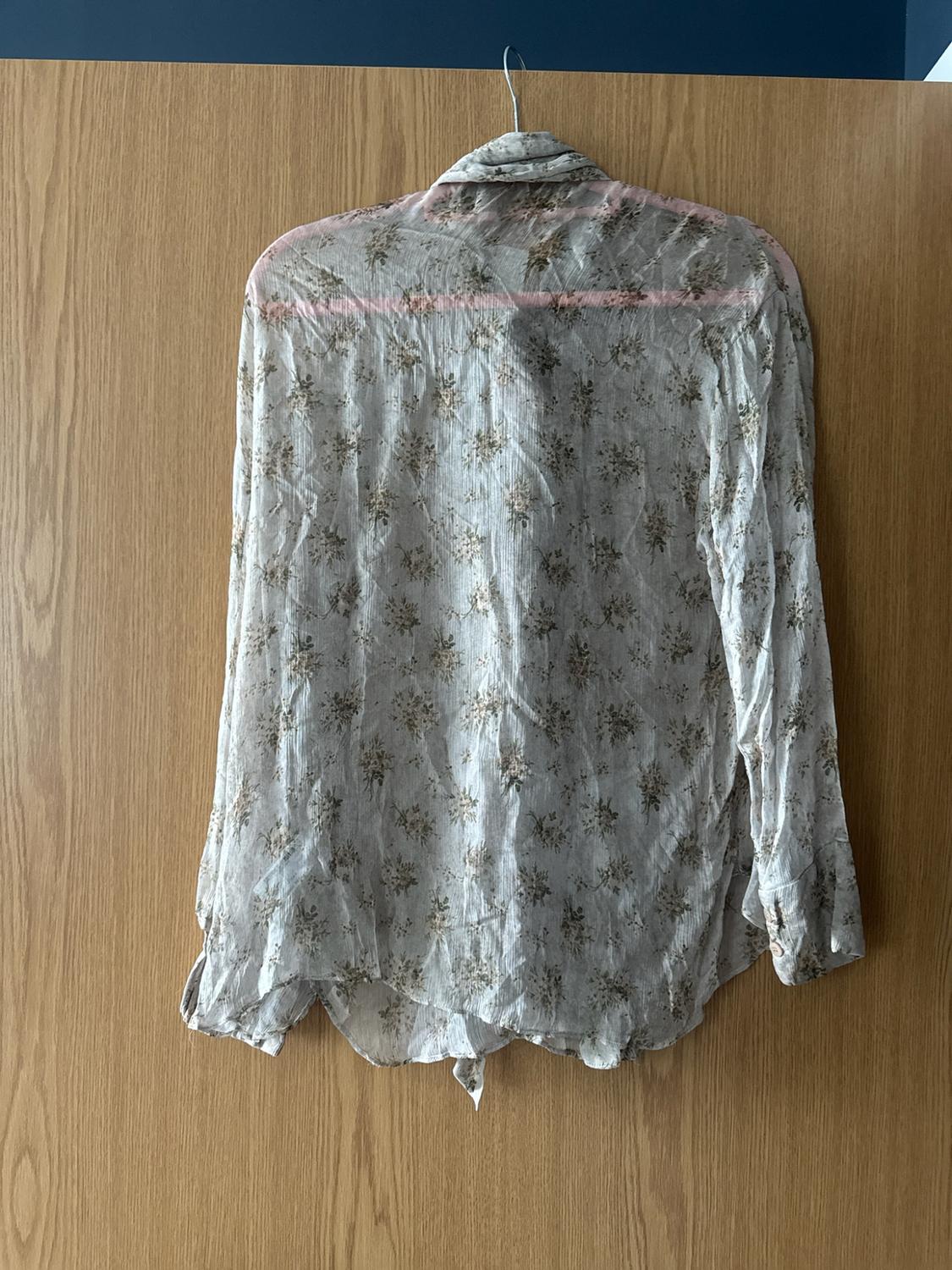 Semi-sheer long-sleeved shirt with a delicate floral print, hanging on a pink coat hanger against a light wood background.