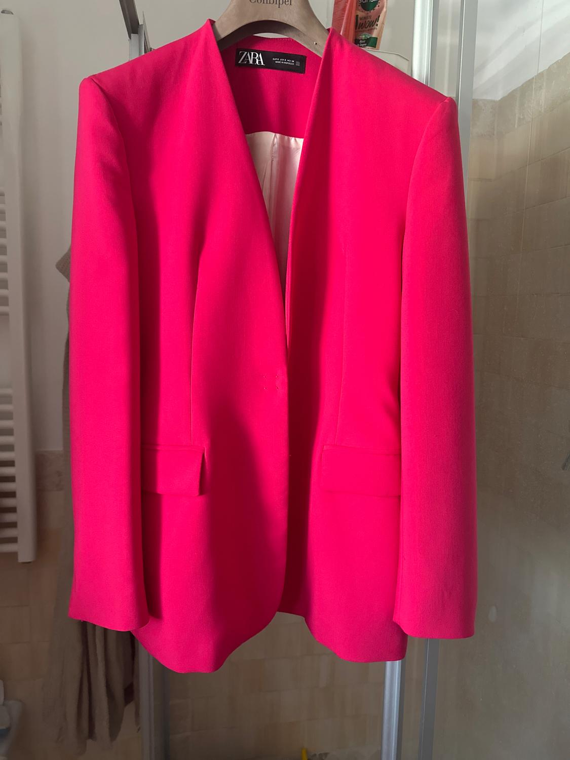 Fuchsia jacket hanging on a wooden hanger with a visible Zara tag, bathroom background with beige tiles and towels.