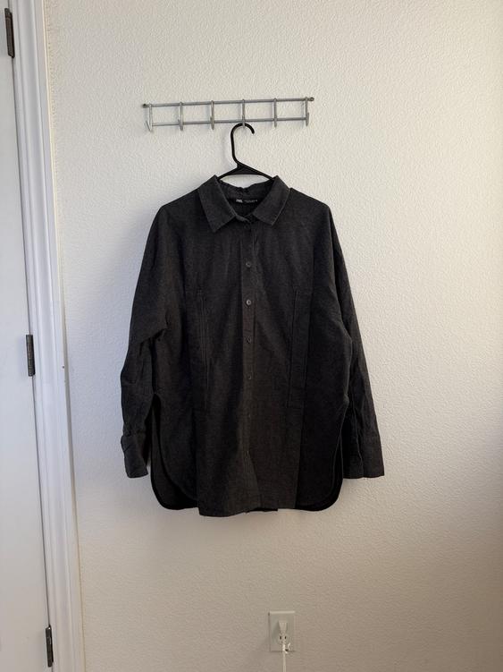 Dark gray shirt hanging on a black hanger against a white wall. The tag says "Zara." In the background, an outlet and a white door.