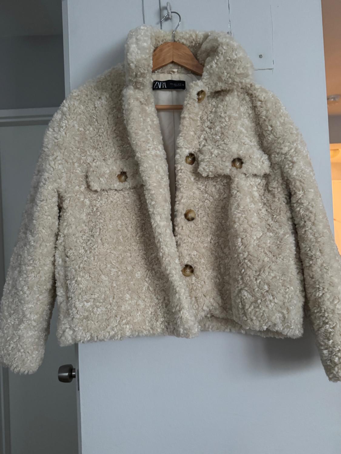Beige fleece jacket hanging on a wooden hanger. It has large buttons and two front flap pockets. Tag "ZV1".