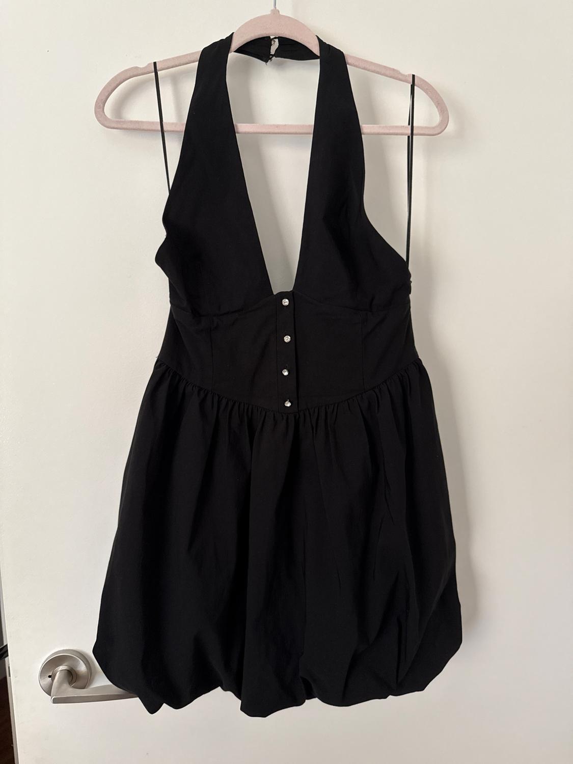 Black halter-neck dress, V-neck, with buttons and a puffball skirt, hanging on a hanger over a white door with a handle.