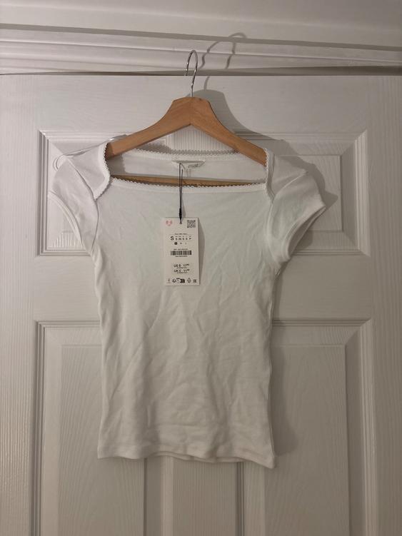 White short-sleeved T-shirt hanging on a wooden hanger over a white door. Visible tag with a price of 12 USD.