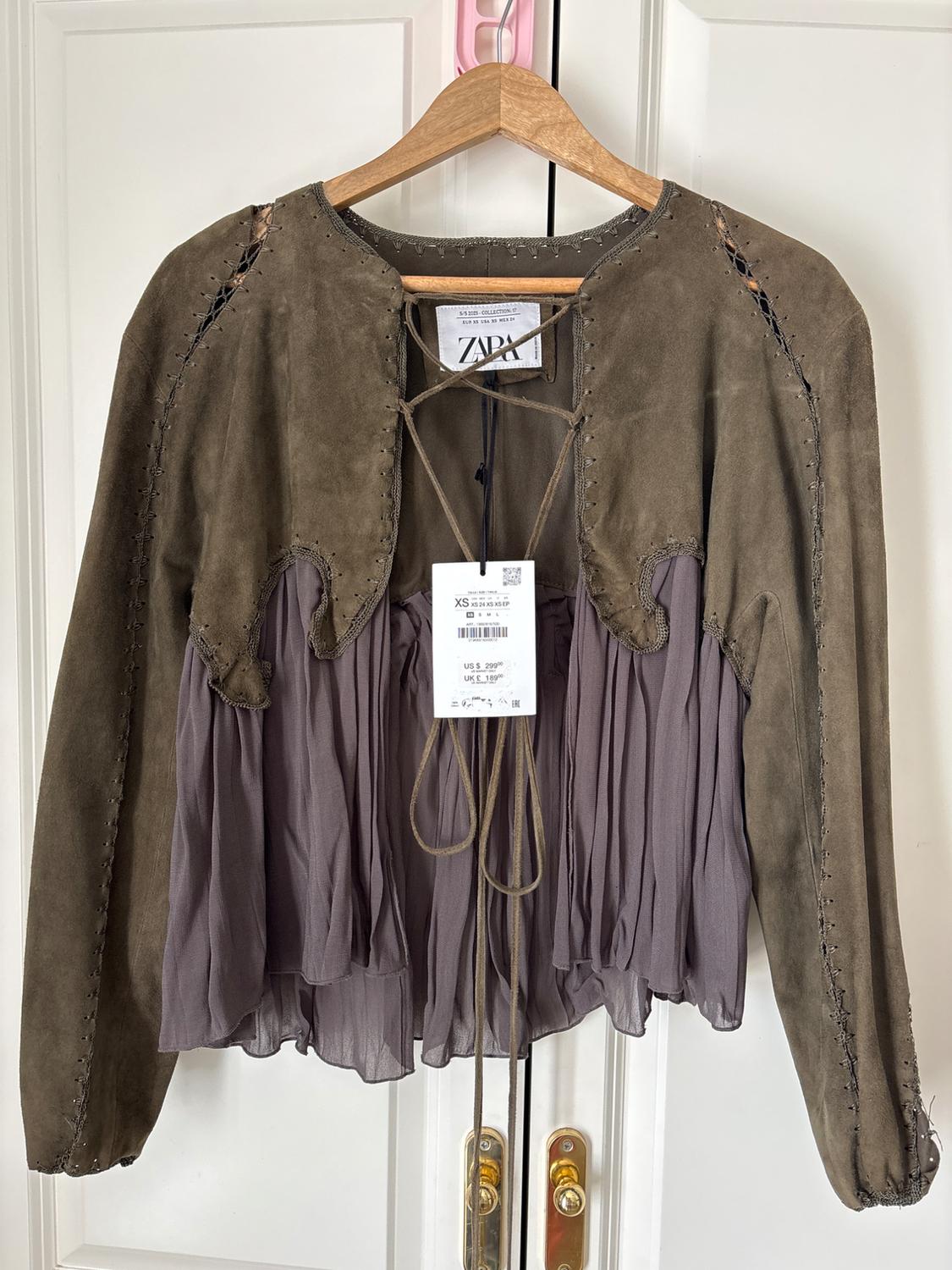 Brown bohemian long-sleeved top, with a lilac pleated hem and lace-up details, hanging on a white door. Visible text: ZARA, XS.