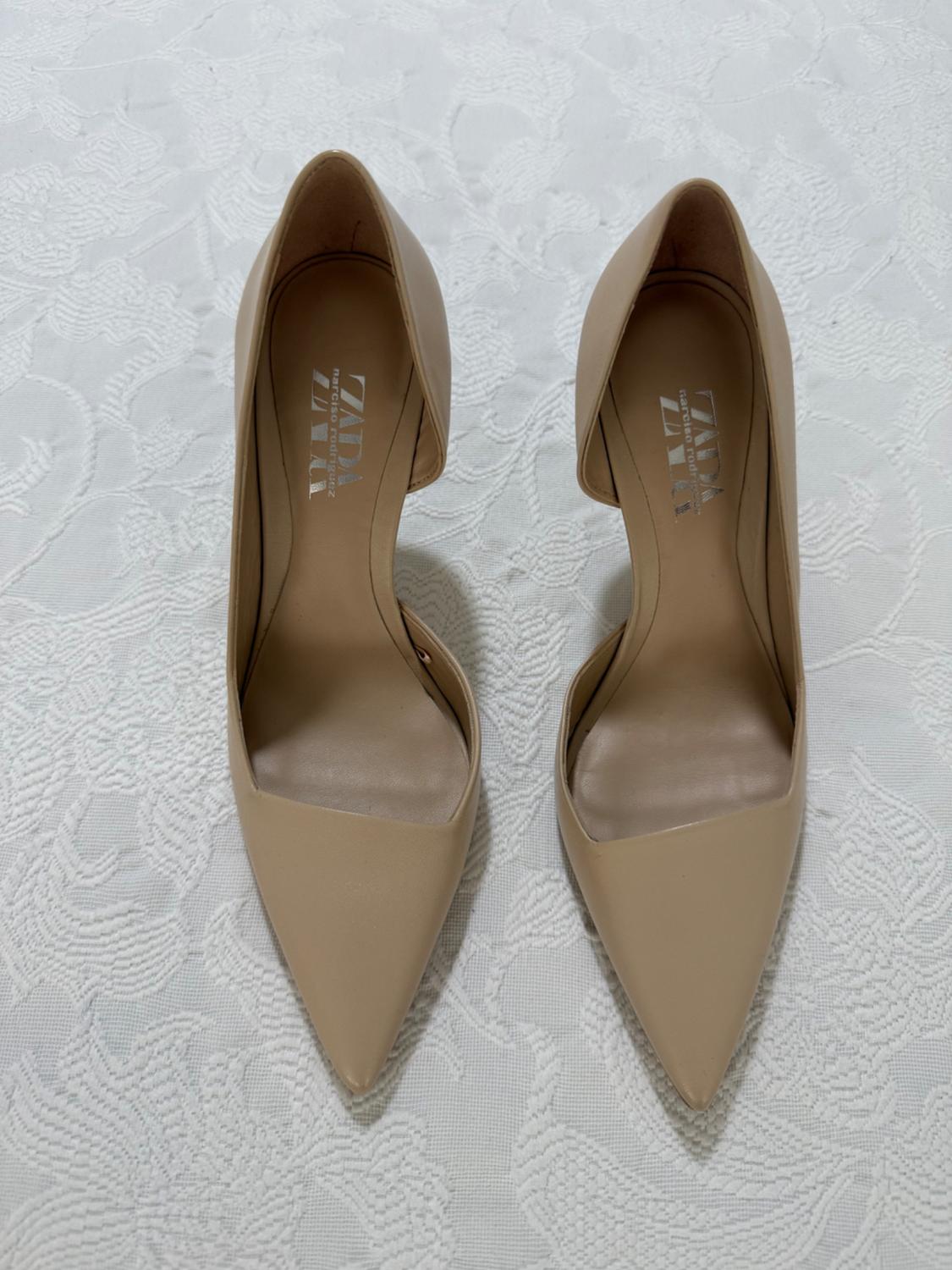 Beige pointed high heels on a textured white fabric background. Visible text: "ZARA".