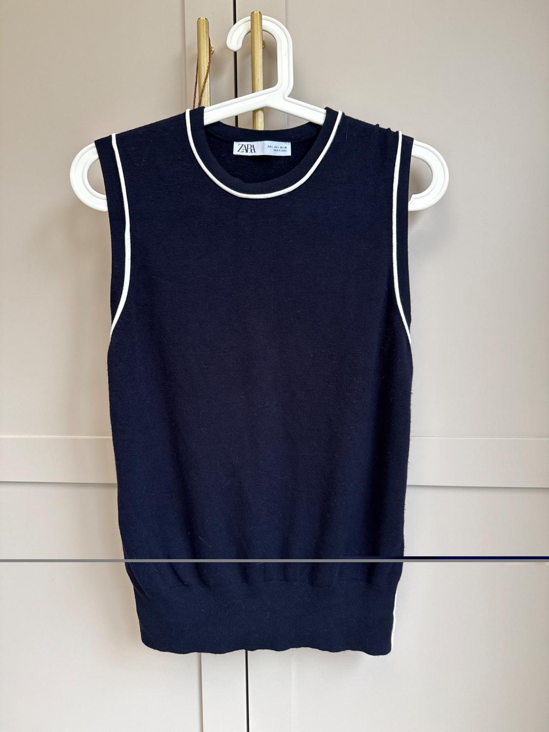 Navy sleeveless waistcoat with white trim hanging on a white hanger against a beige background. Visible tag: ZARA.