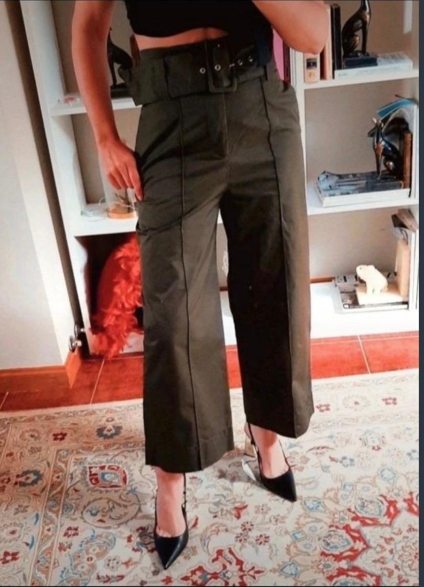 Woman in a black top and high-waisted green trousers, posing in a room with a bookshelf and decorative rug. She is wearing black heels.