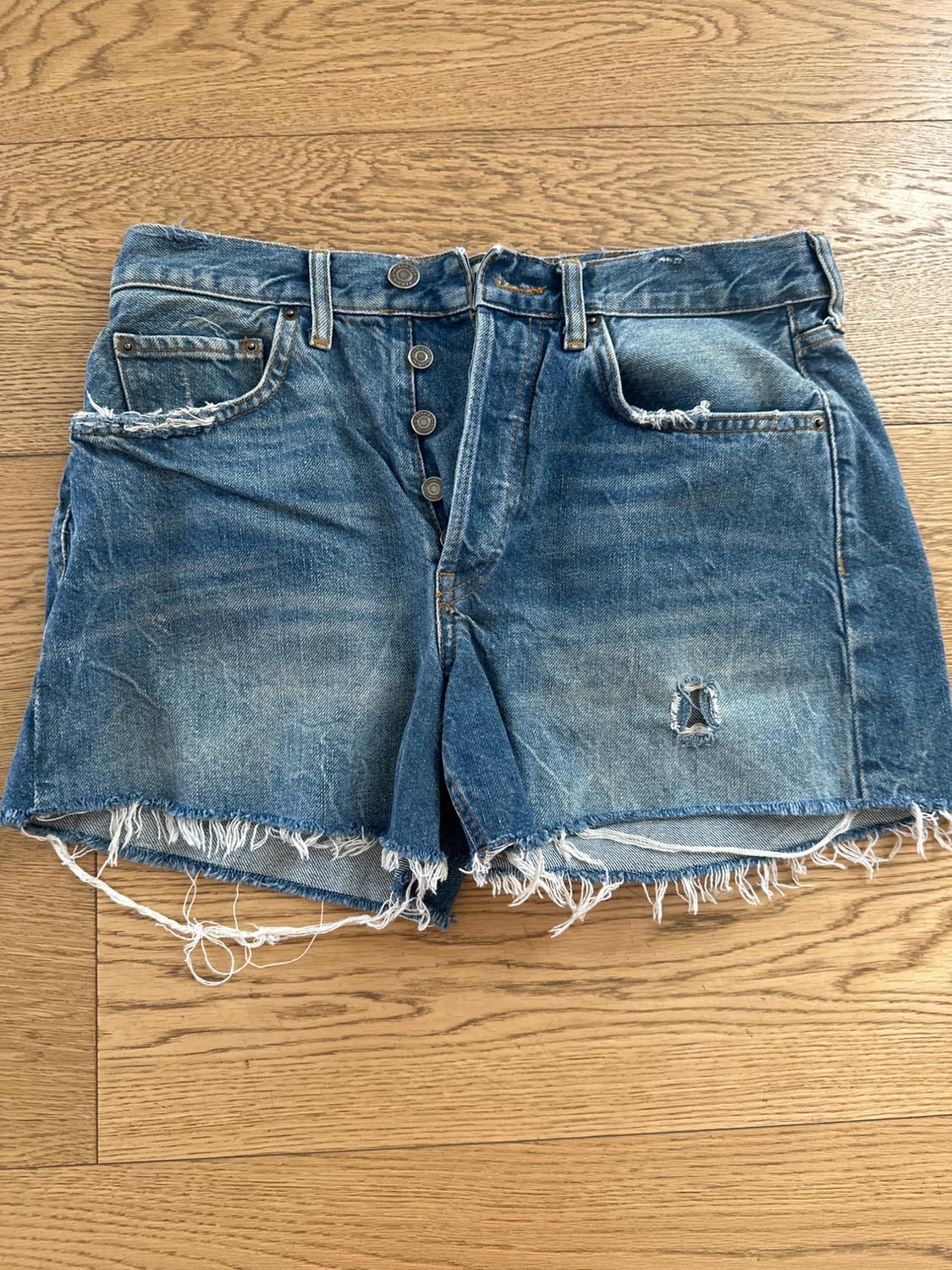 Distressed denim shorts with front buttons and frayed edges on a light wooden floor.