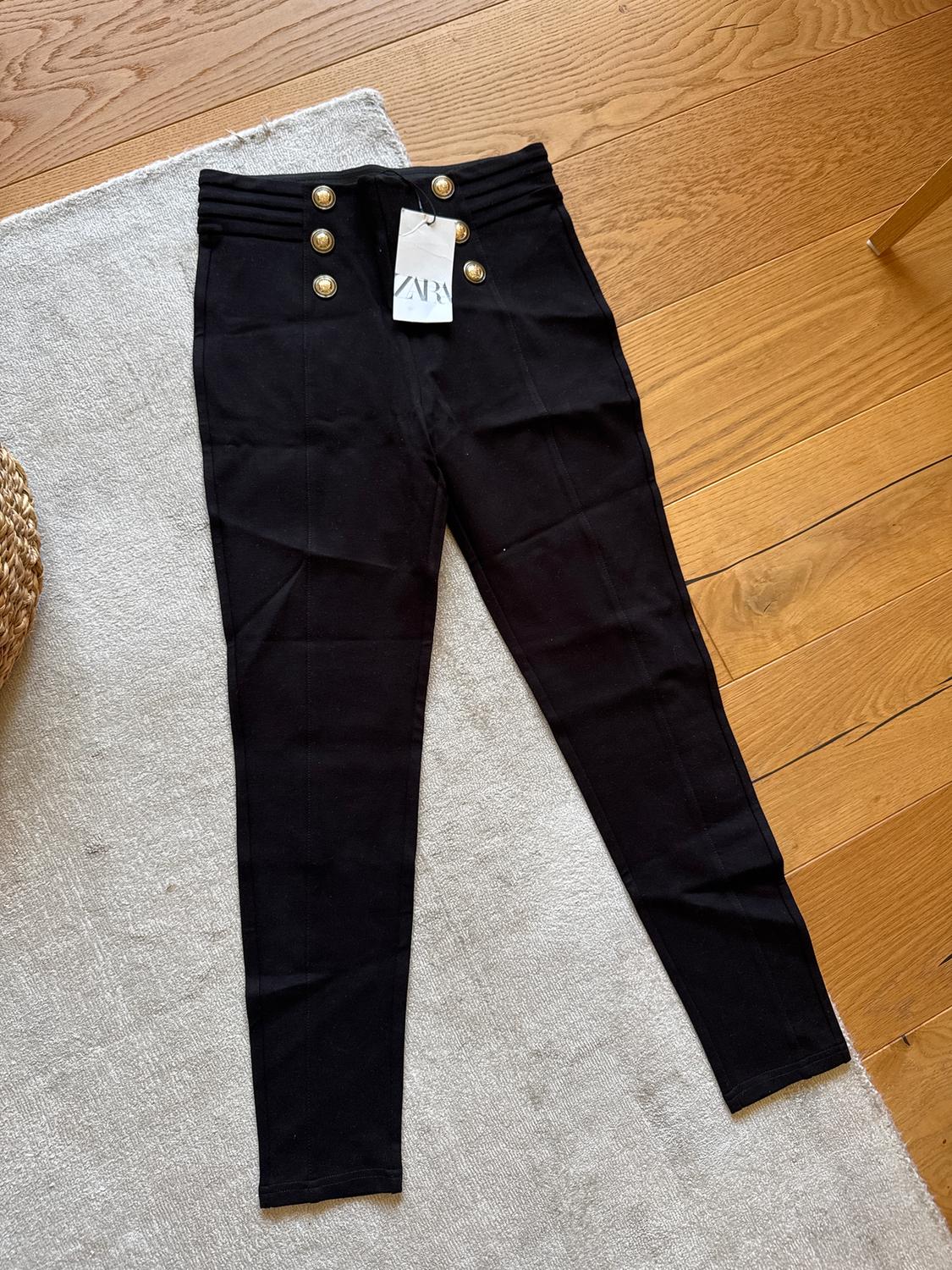Black trousers with gold buttons on the waist, visible Zara tag, on a light rug and wooden floor.