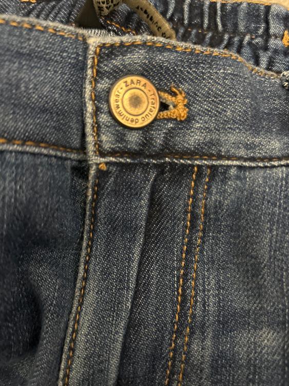 Detail of blue denim trousers with a metal button that says "ZARA TRAFALGAR denimwear", visible orange thread stitching.