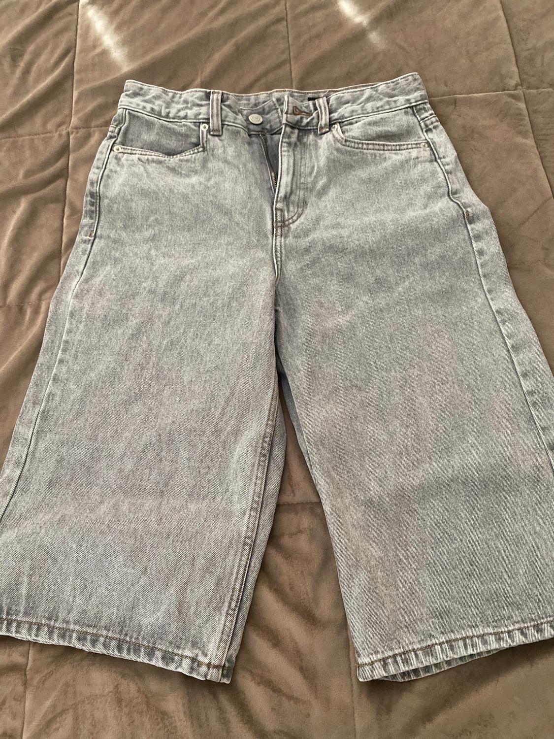 Light-wash, wide-leg cowboy Bermuda shorts, with the zip partly open, on a textured brown quilt in a casual style.
