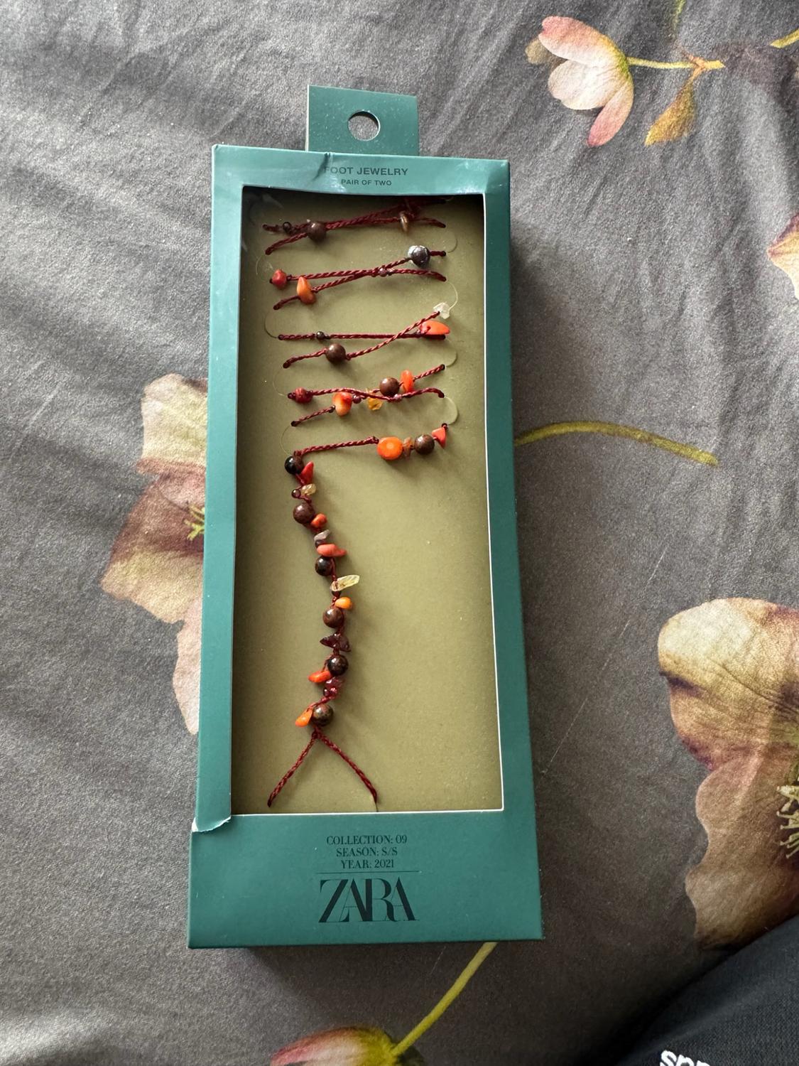 Green box on floral fabric with red thread and bead jewellery. Text: FOOT JEWELRY PAIR OF TWO COLLECTION: 09 SEASON: S/S YEAR: 2021 ZARA.