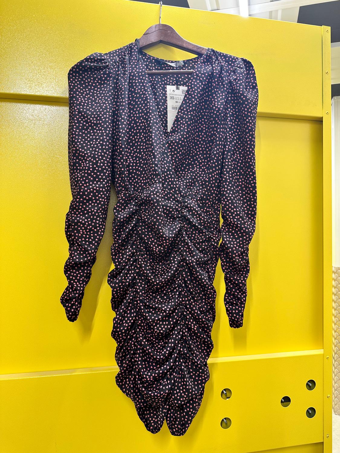Black dress with a white and red heart print, long sleeves and ruching, hanging on a hanger against a yellow background.