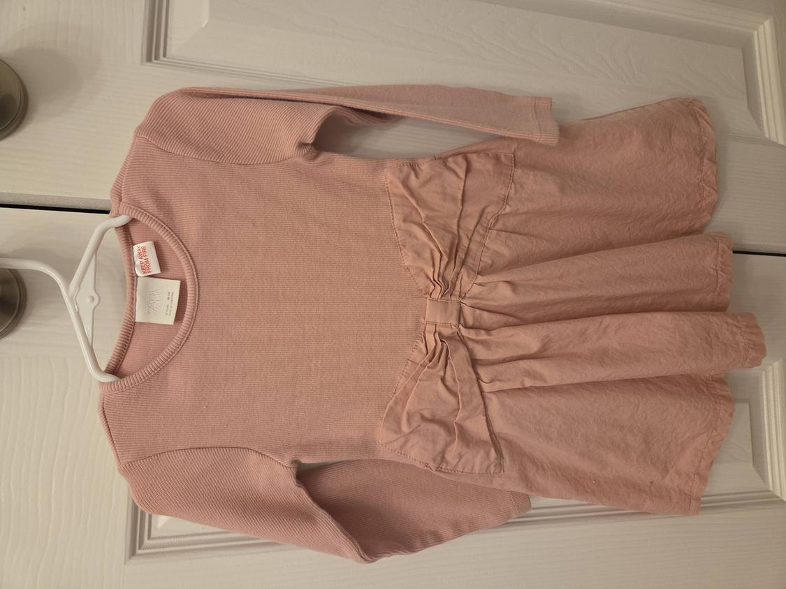Light pink, long-sleeved dress with a bow at the waist, hanging on a white hanger over a white door.