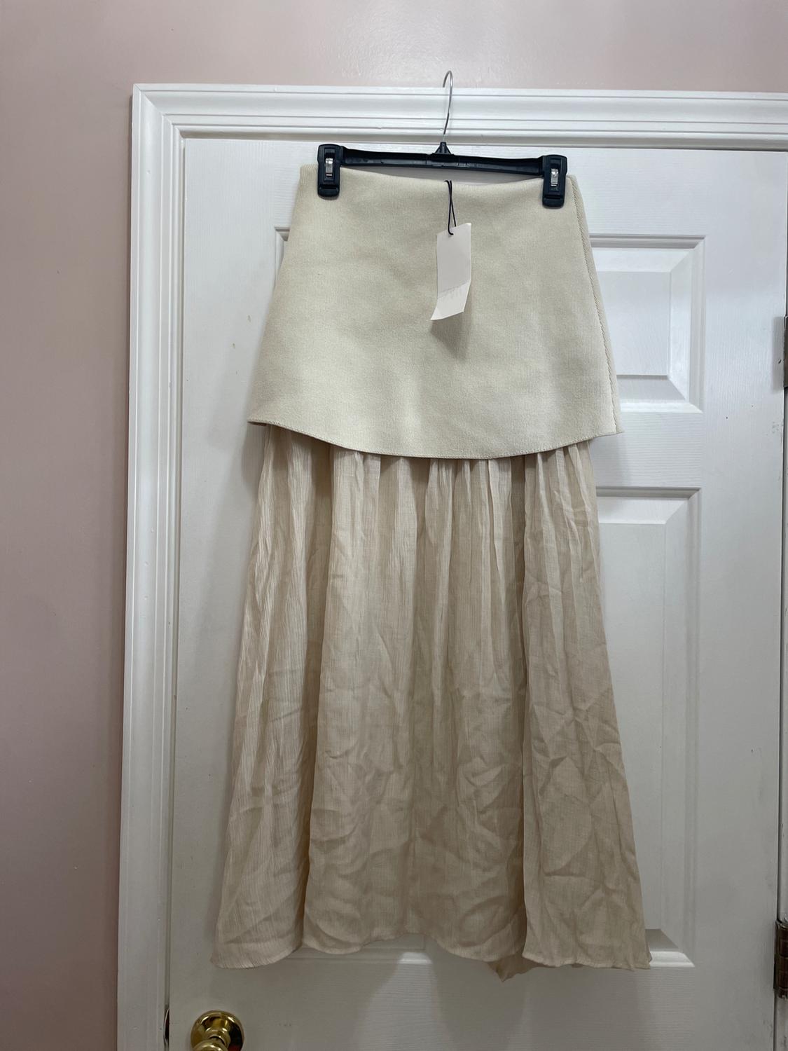 Cream-colored long skirt with a tag, hanging on a white door. Double-layer design: short and smooth top layer, flowing and wrinkled bottom layer.