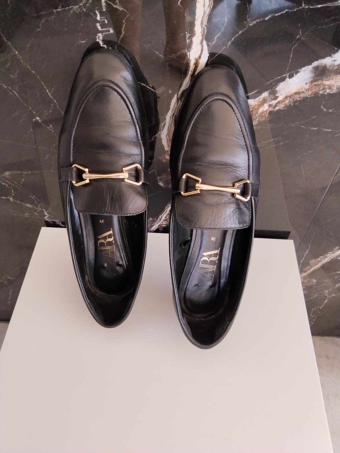 Black loafers with a gold-tone metallic detail on a white surface against a dark marble background. Visible text: ZARA 39.