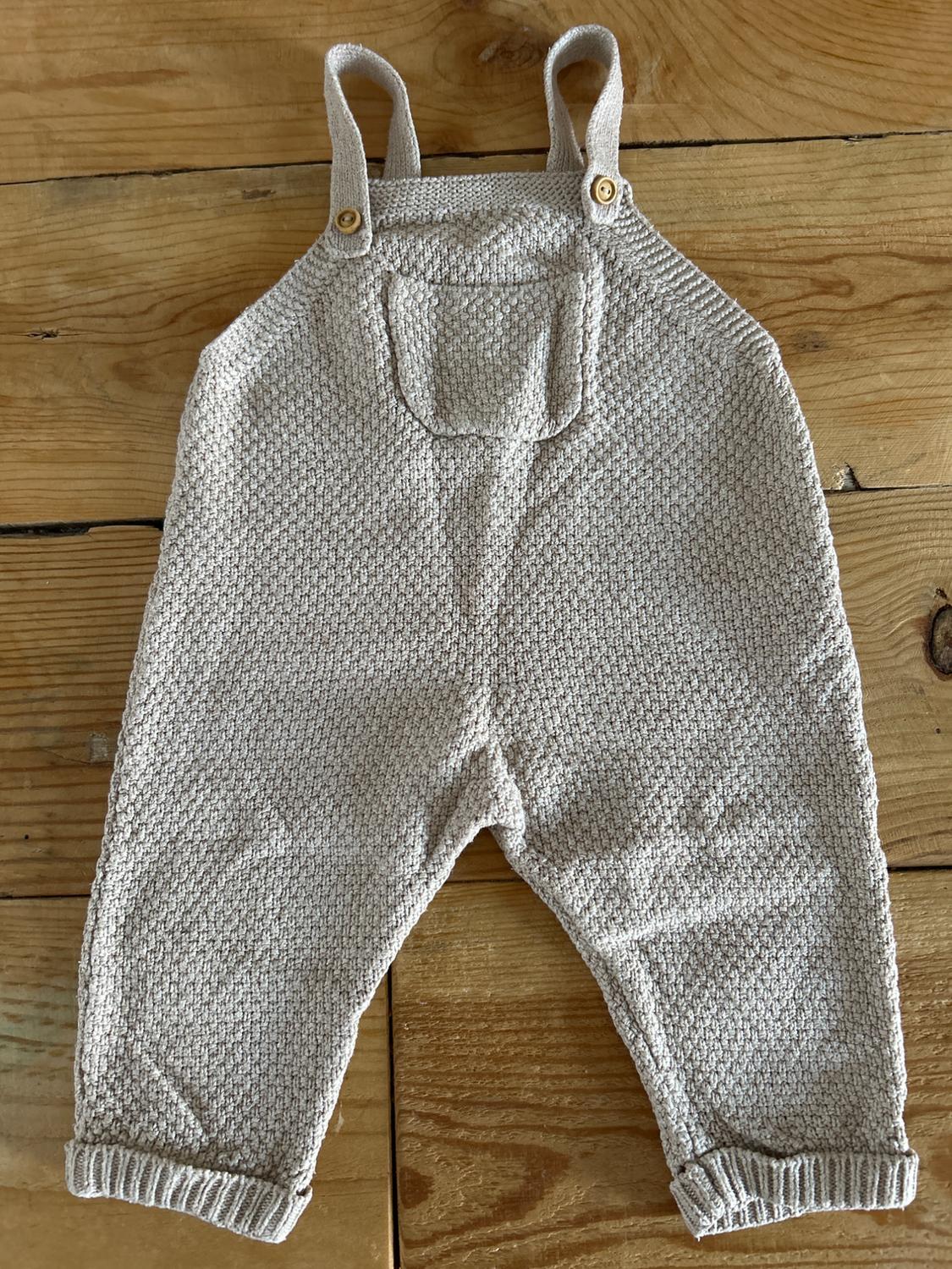 Knitted baby dungarees in beige with straps and wooden buttons, on a light wooden background.