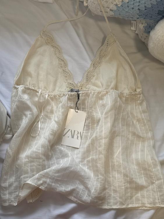 White camisole with lace on the neckline, on a bed. Visible tag with the text "ZARA".