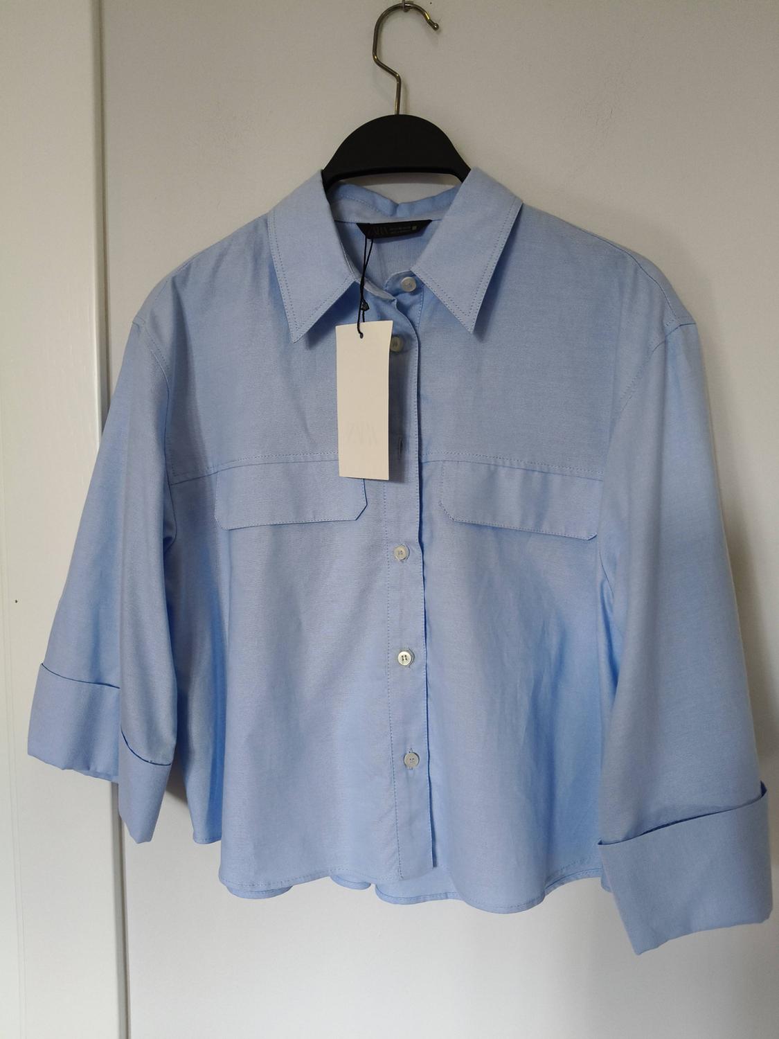Light blue, long-sleeved shirt hanging on a black hanger, with a white label visible on the collar.