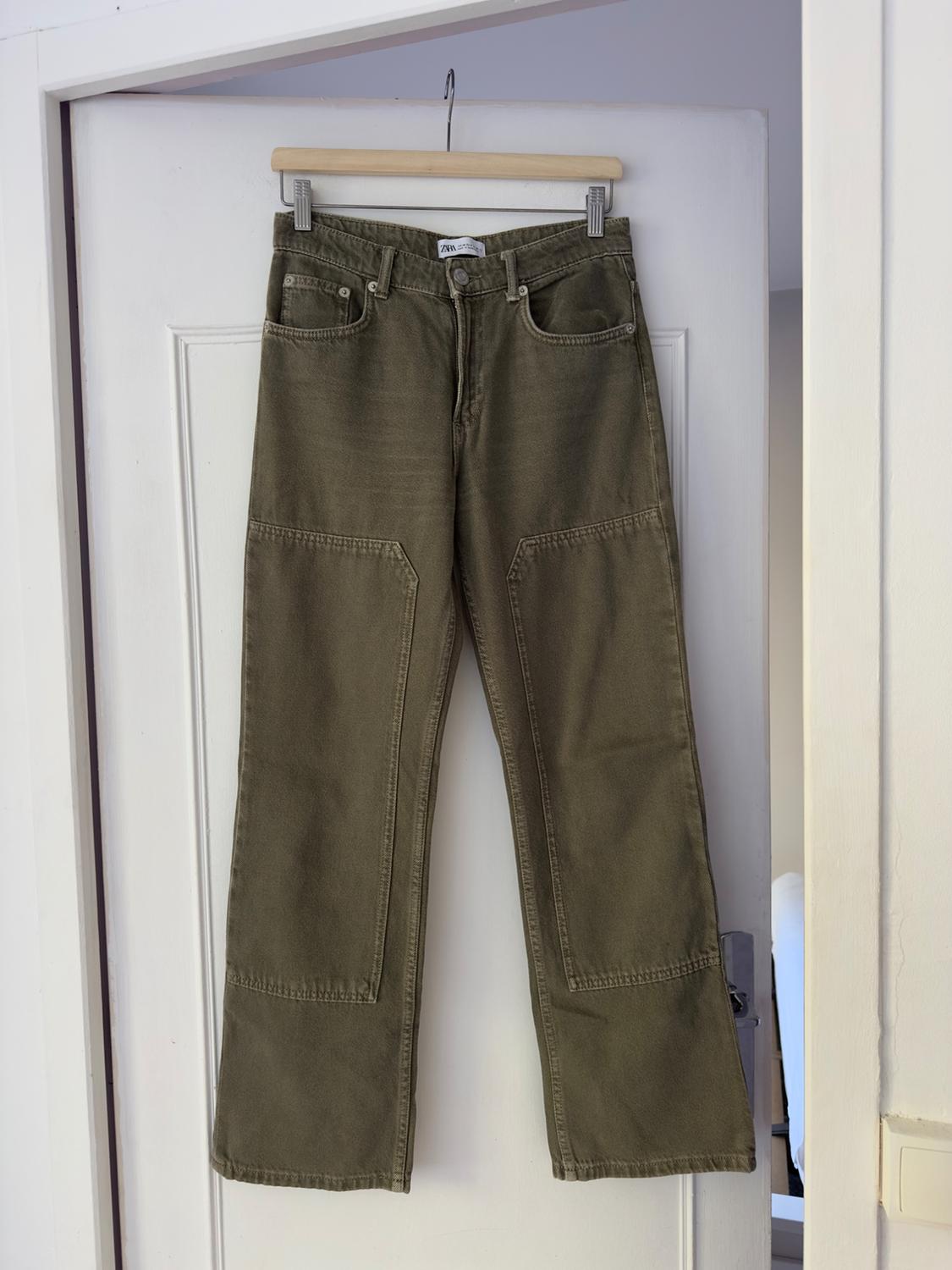 Green trousers hung on a wooden hanger over a white door. Front pockets and visible seams.