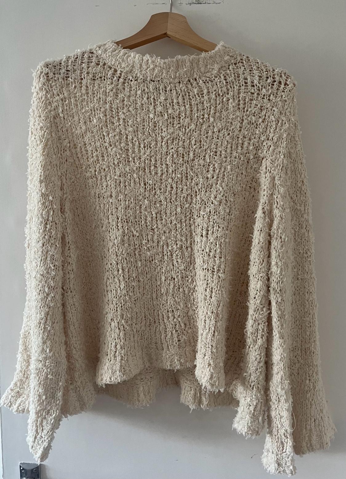 Cream-coloured long-sleeved loose-fit textured knit jumper, hanging on a wooden hanger against a white wall.