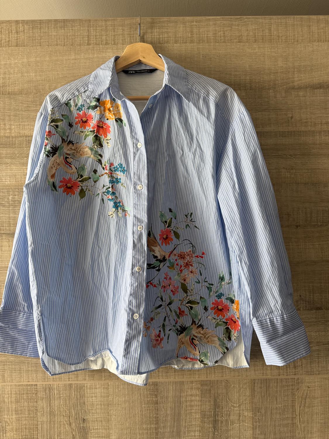 Blue striped shirt with a floral and bird print in red, orange, and green tones, hanging on a wooden hanger.