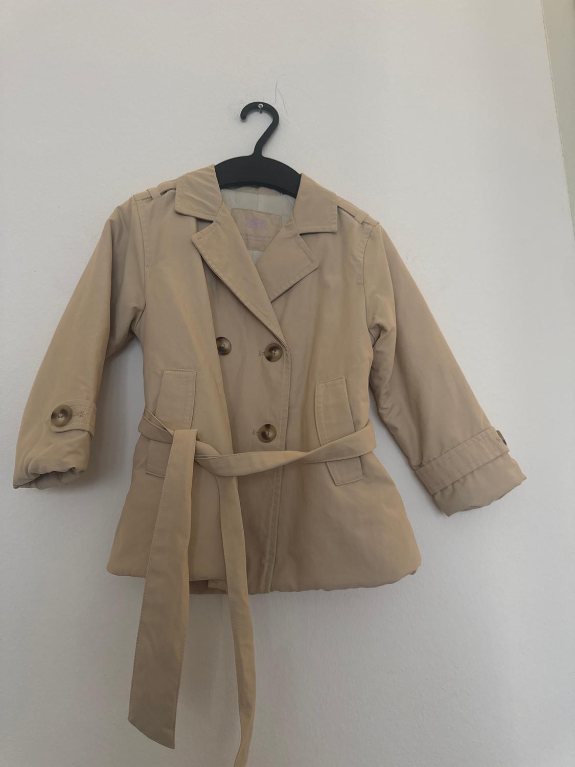 Kids' beige coat hanging on a black hanger against a white wall. Double-breasted design with a belt.