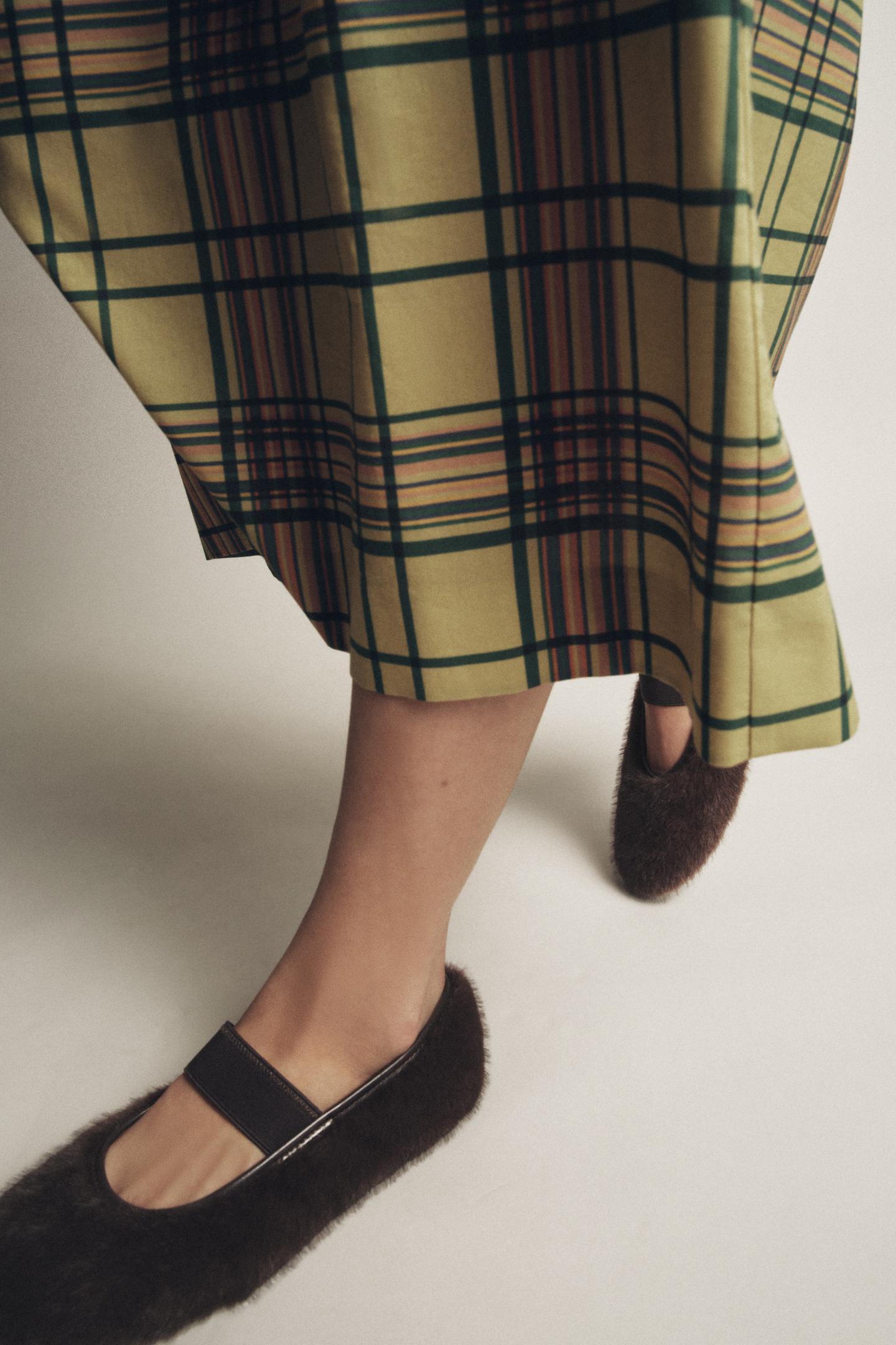 Close-up of chocolate brown flats with elastic strap and plaid print skirt in shades of green and yellow.