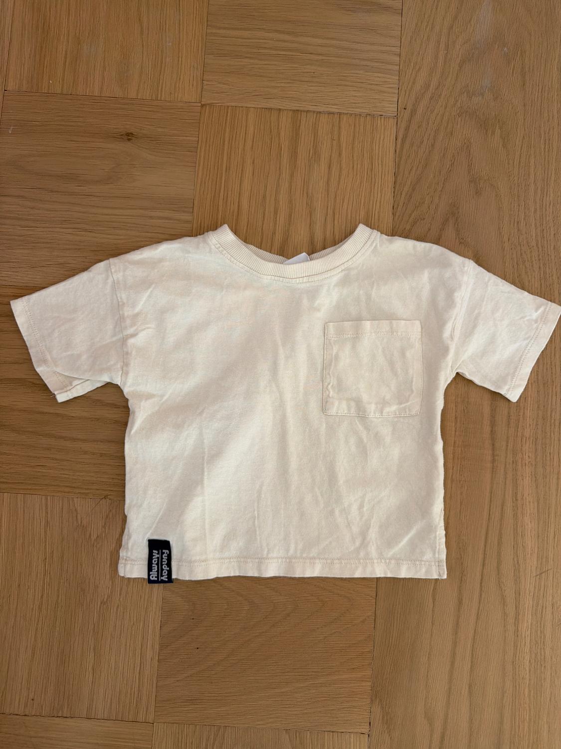 White short-sleeved T-shirt with chest pocket, on a wooden floor. Black label with "Funday Monday" text on the hem.