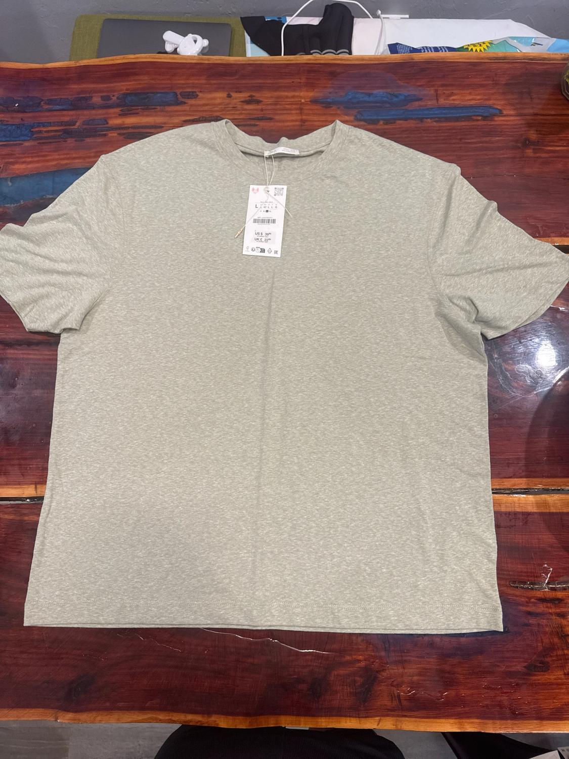 Light green heather short-sleeved t-shirt with its tag attached, laid out flat on a wooden table with blue resin inlay.
