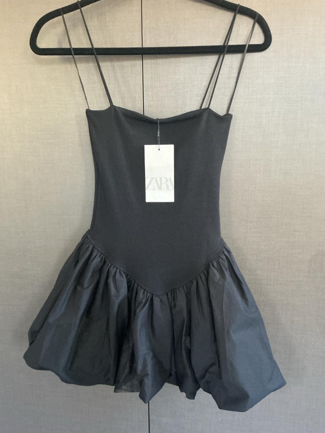 Short black spaghetti strap dress, fitted bodice and bubble skirt hanging on a hanger. Label with the text Zara.