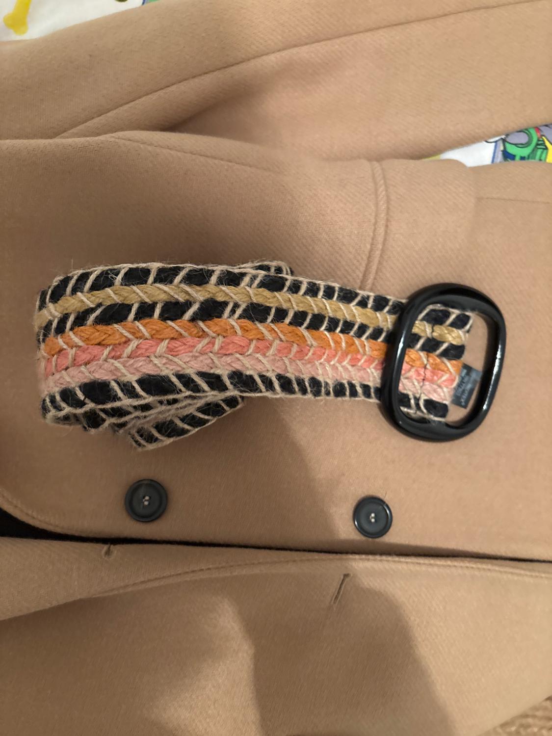 Detail of a beige coat with dark buttons and a multicoloured braided belt with a black buckle on a light surface.