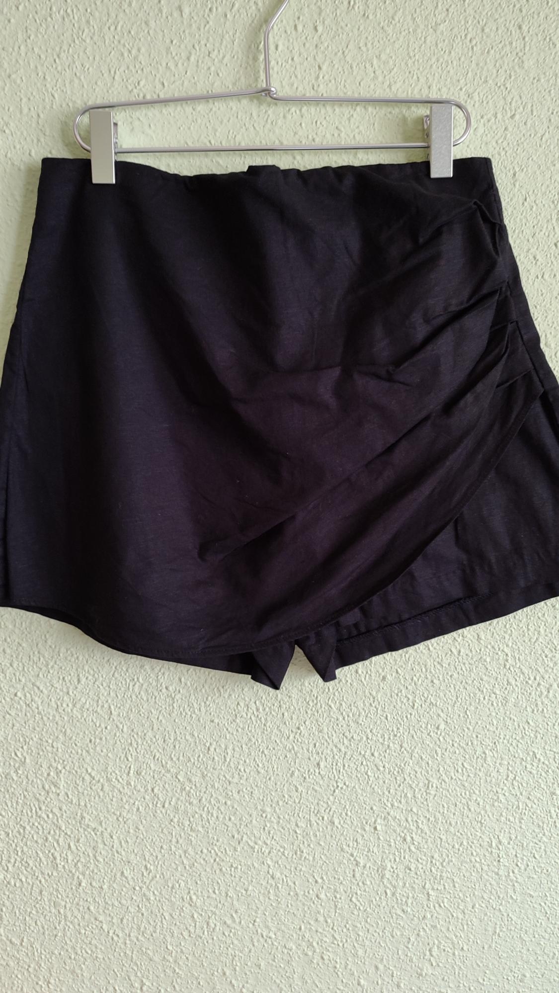 Black culottes with draped front detail, hanging on a metal hanger against a light, textured wall.