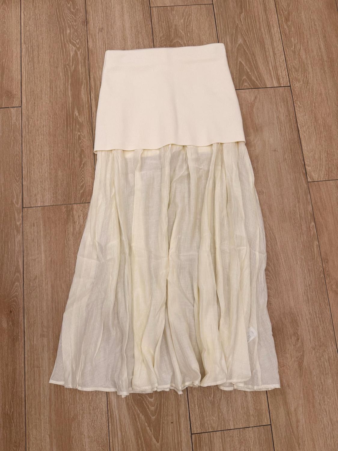 Long cream-coloured skirt on a wooden floor. Design with a plain upper section and a flowing, semi-transparent fabric lower section.