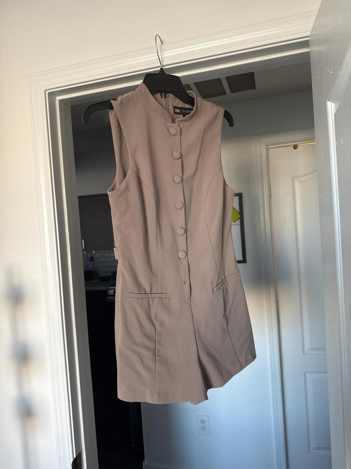 Beige high-neck short jumpsuit with front buttons hanging on a white door indoors. Tag with the text ZARA.