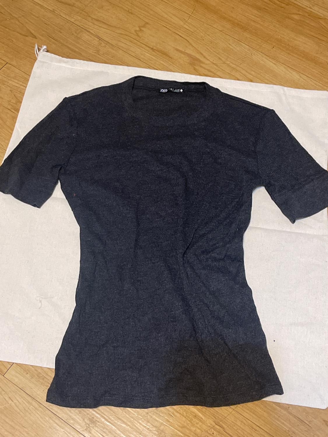 Black short-sleeved t-shirt on white fabric on wooden floor. Visible tag on the collar.