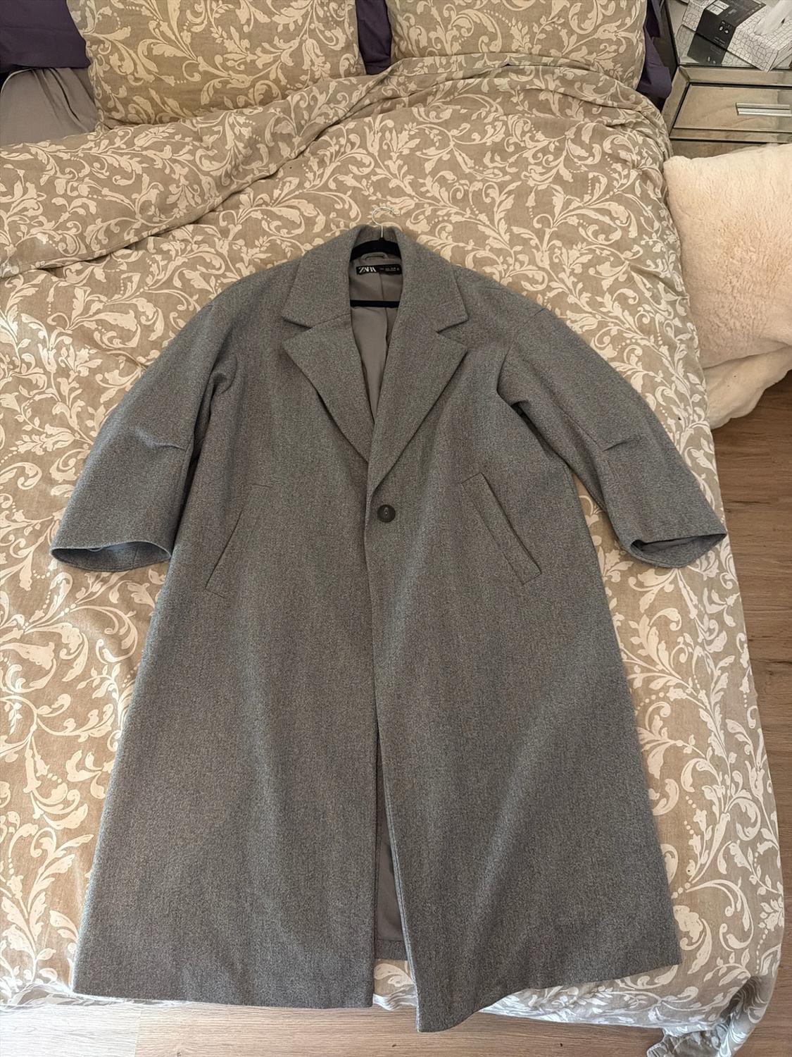 Long grey ZARA coat with wide lapels and one button, laid out on a bed with a beige patterned duvet.